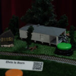 Diorama with 3D printed shack, trees, grave, braille & large print labels, QR code and touch button