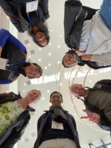 six people smiling down at the camera, with a white ceiling several stories above them
