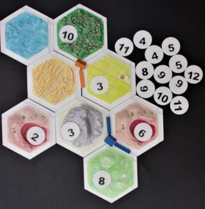Catan game pieces: hexagonal tiles with 3D terrain, discs with numbers in print and braille, and town/road pieces