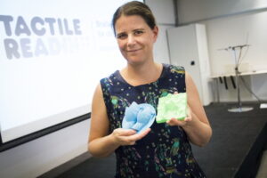 Leona Holloway holding two 3D prints on stage with "tactile reading" on the display screen