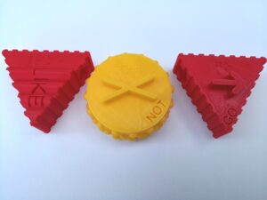 Three 3D printed pieces with print, braille and a tactile symbol. "Like" and "Go" are red triangles with ridges on the side. "Not" is a yellow circle with half spheres on the side. 